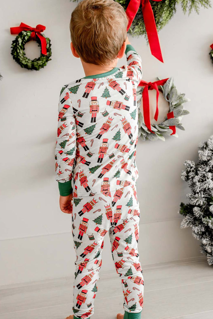 Back view of boy wearing Caden Lane's white toy soldier print pajamas, featuring nutcrackers and Christmas trees, with green cuffs, reaching toward a wreath.