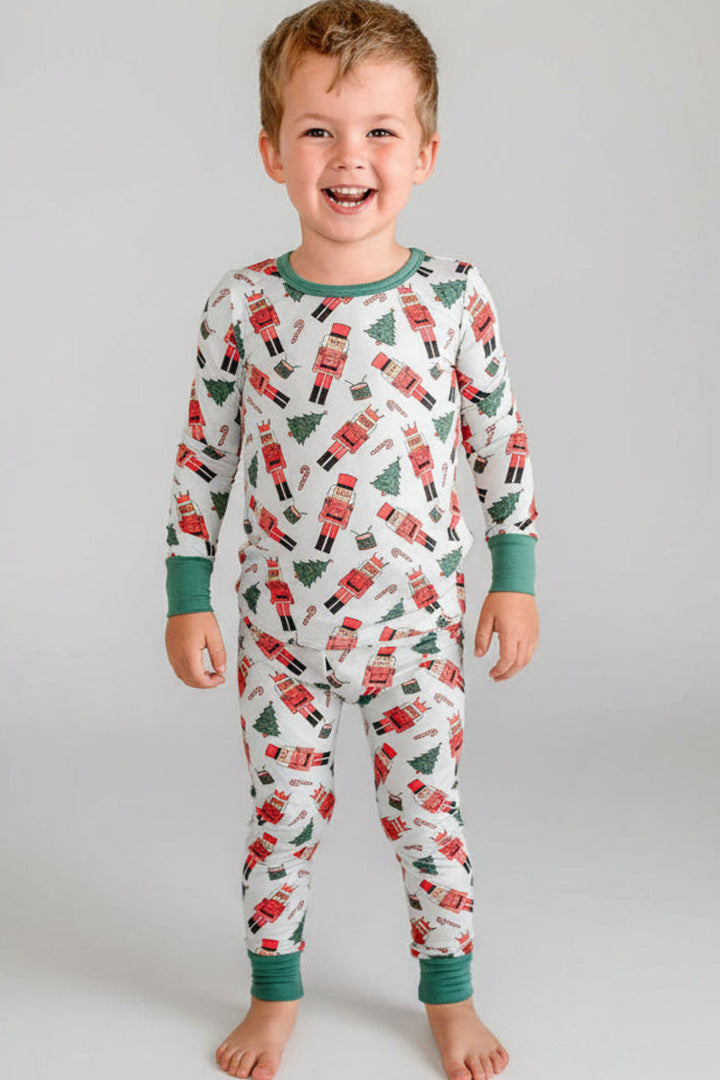 Boy smiling while wearing Caden Lane's white toy soldier print pajamas, featuring nutcrackers and Christmas trees, with green cuffs.