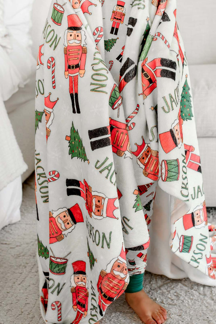 Boy smiling while wrapped in Caden Lane's red toy soldier print blanket personalized with the name "Jackson," featuring nutcrackers, Christmas trees, and candy canes.