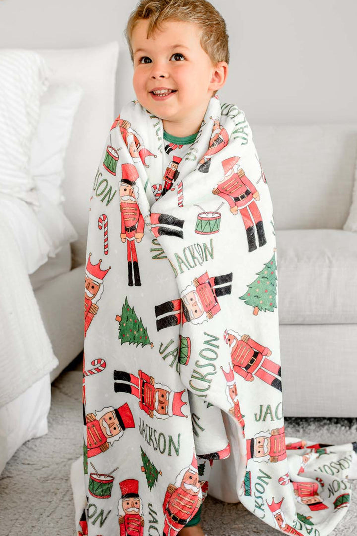 Close-up of Caden Lane's red toy soldier print blanket draped over a child, personalized with the name "Jackson," featuring nutcrackers, Christmas trees, and candy canes.