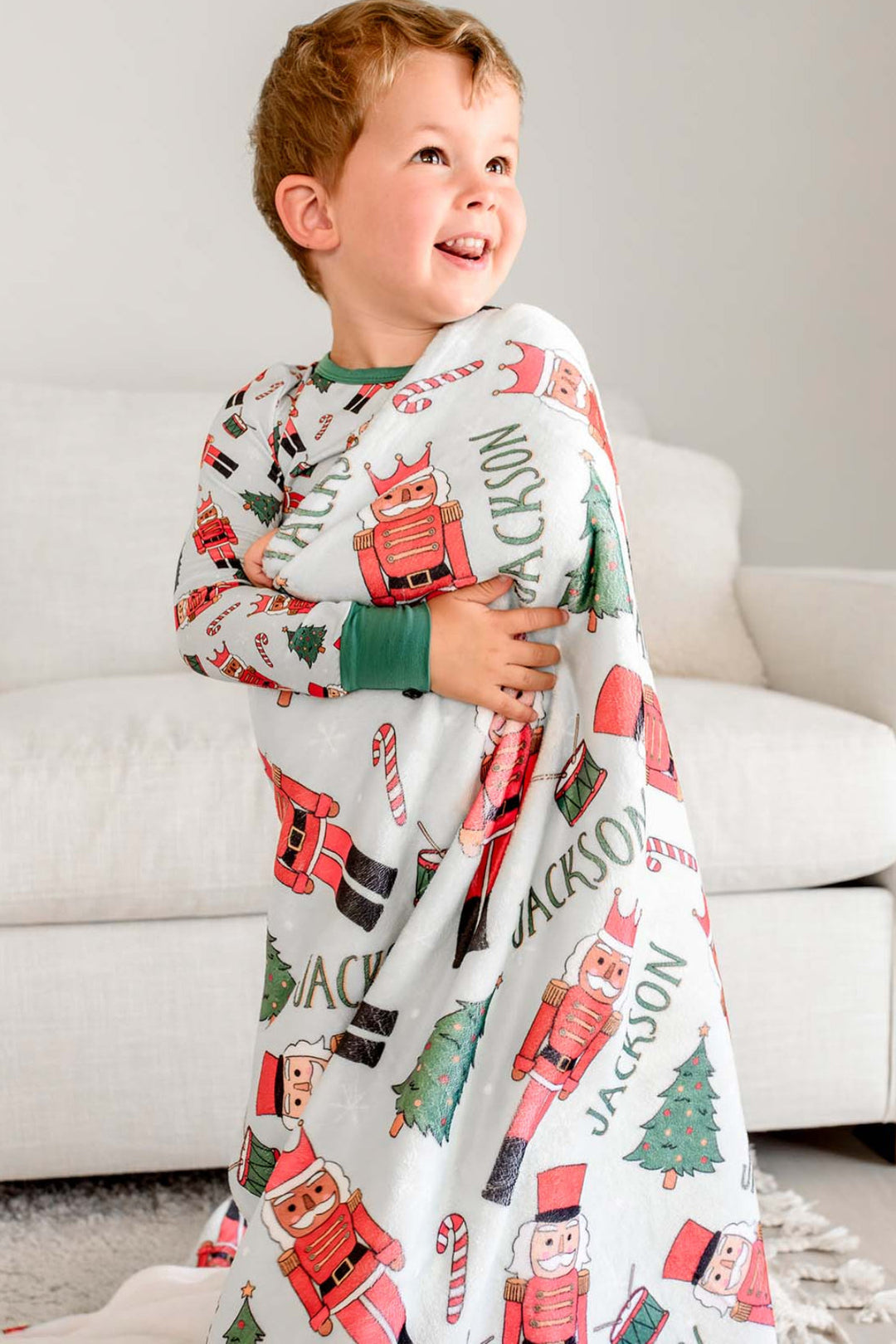 Boy smiling while hugging Caden Lane's red toy soldier print blanket personalized with the name "Jackson," featuring nutcrackers, Christmas trees, and candy canes.