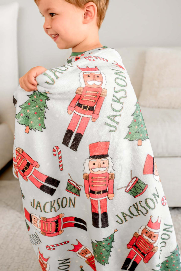 Close-up of a boy wearing Caden Lane's blanket over his shoulder, personalized with the name "Jackson," featuring nutcrackers, Christmas trees, and candy canes.