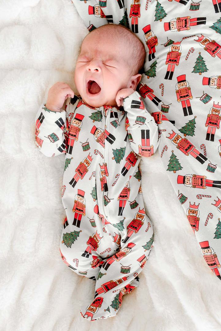 Newborn boy yawning while wearing Caden Lane's white toy soldier print zip-up pajamas, featuring nutcrackers and Christmas trees.