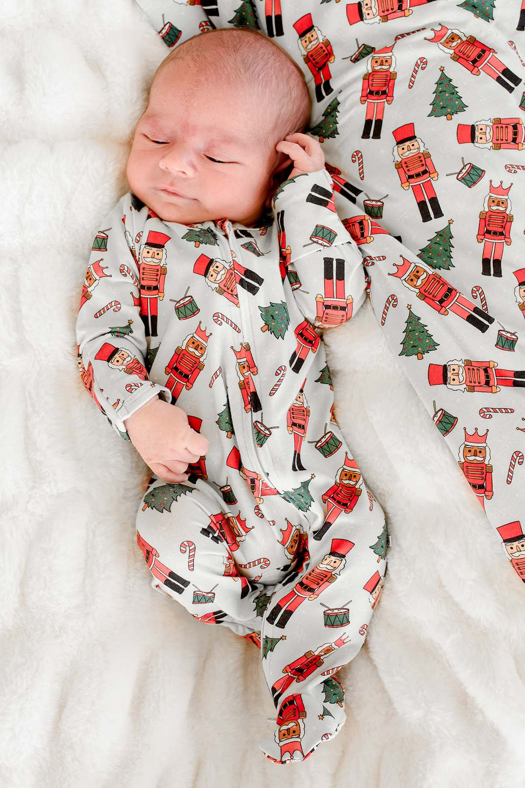 Newborn boy sleeping while wearing Caden Lane's white toy soldier print zip-up pajamas, featuring nutcrackers and Christmas trees.