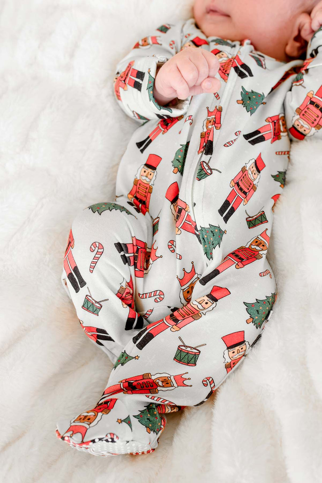 Close-up of newborn boy wearing Caden Lane's white toy soldier print zip-up pajamas, featuring nutcrackers and Christmas trees.