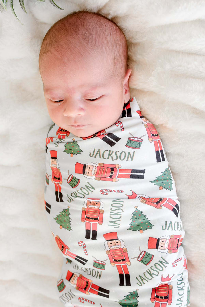 Close-up of newborn boy swaddled in Caden Lane's white toy soldier print blanket personalized with the name "Jackson," featuring nutcrackers, Christmas trees, and candy canes.