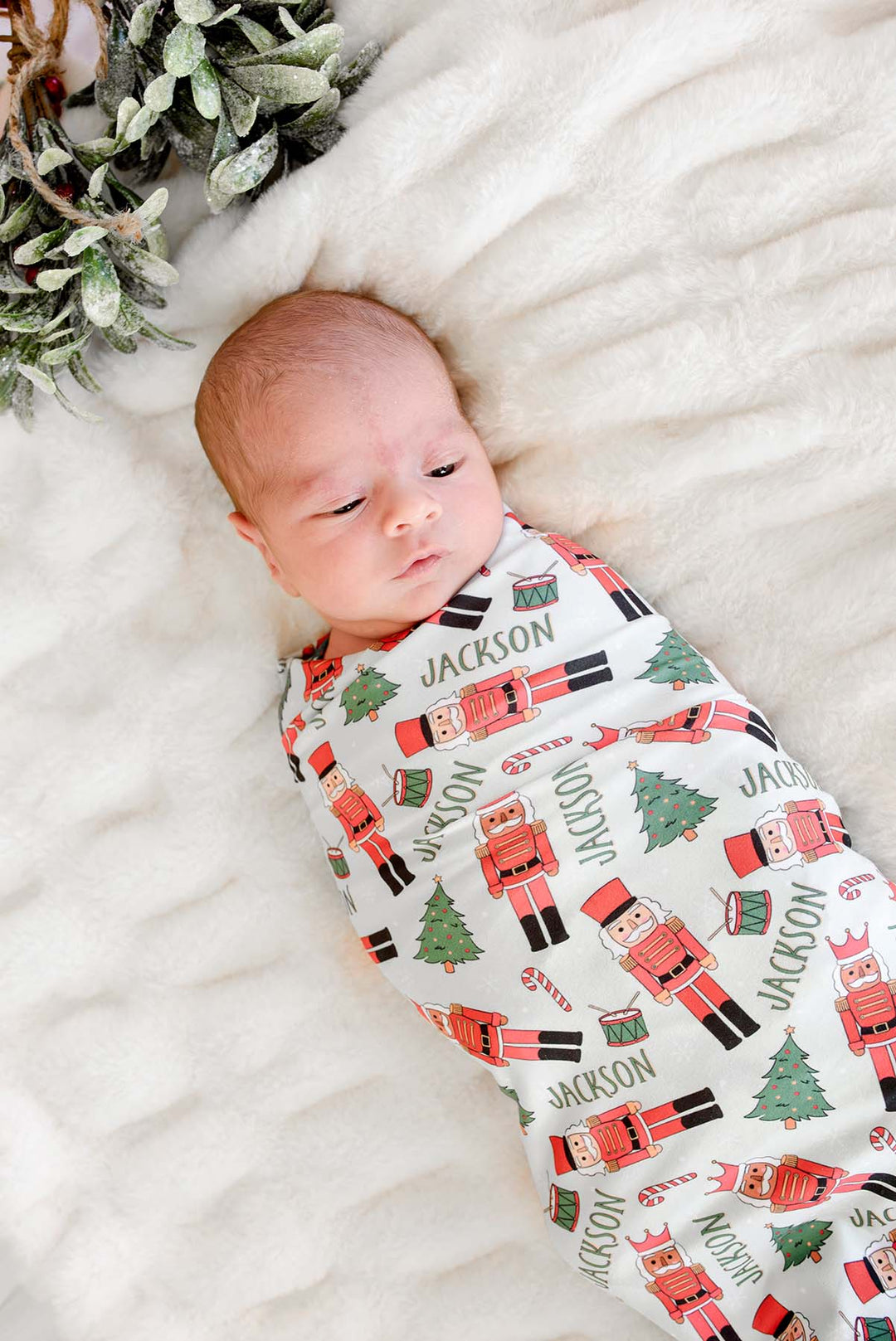 Newborn boy swaddled in Caden Lane's white toy soldier print blanket personalized with the name "Jackson," featuring nutcrackers, Christmas trees, and candy canes, with mistletoe in the background.