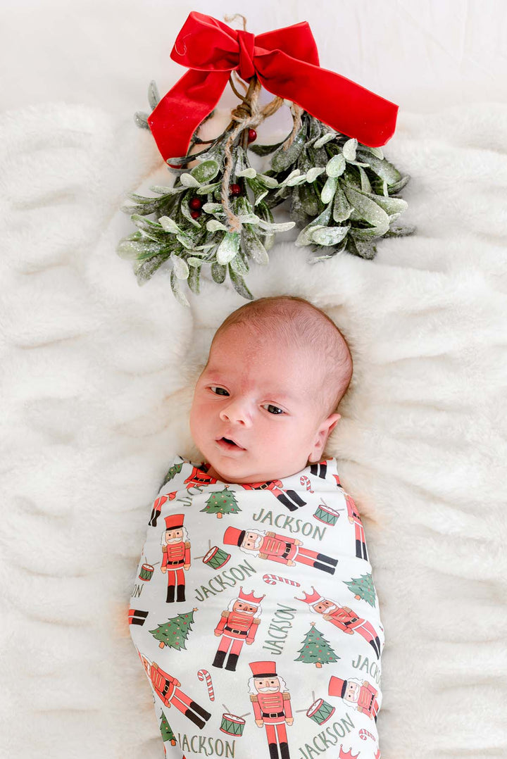 Newborn boy swaddled in Caden Lane's white toy soldier print blanket personalized with the name "Jackson," featuring nutcrackers, Christmas trees, and candy canes, with mistletoe and a red bow above.