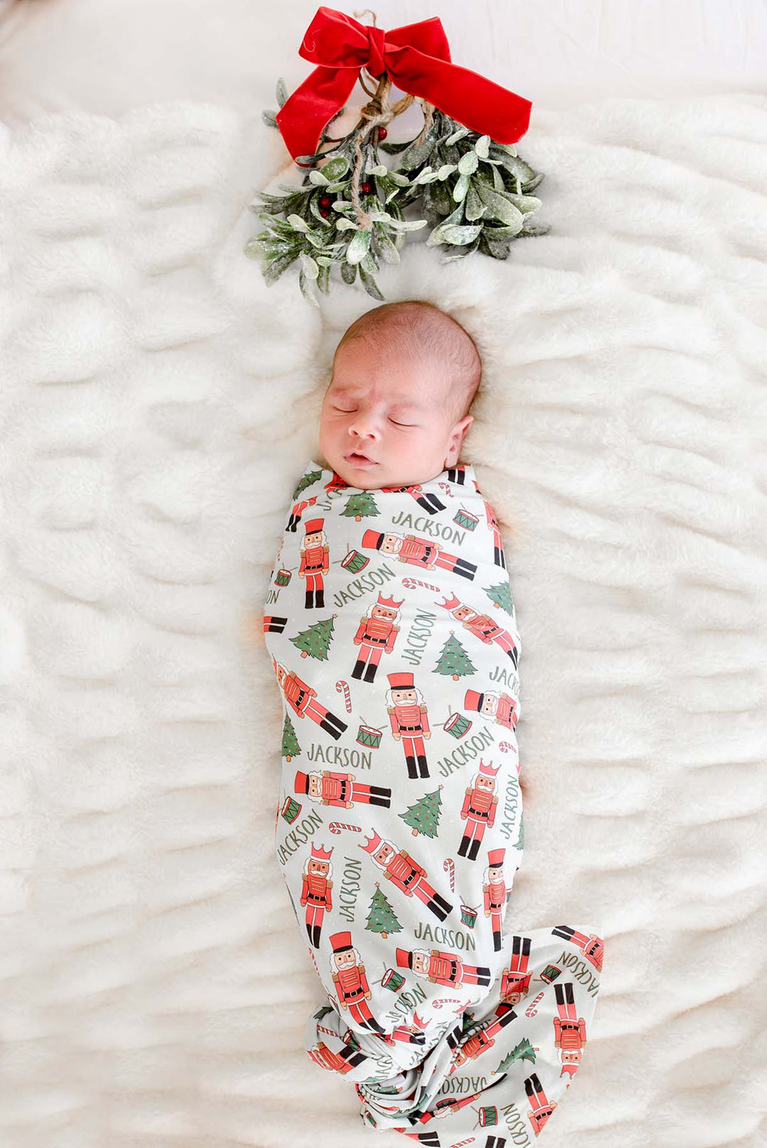 Newborn boy sleeping while swaddled in Caden Lane's white toy soldier print blanket personalized with the name "Jackson," featuring nutcrackers, Christmas trees, and candy canes, with mistletoe and a red bow above.