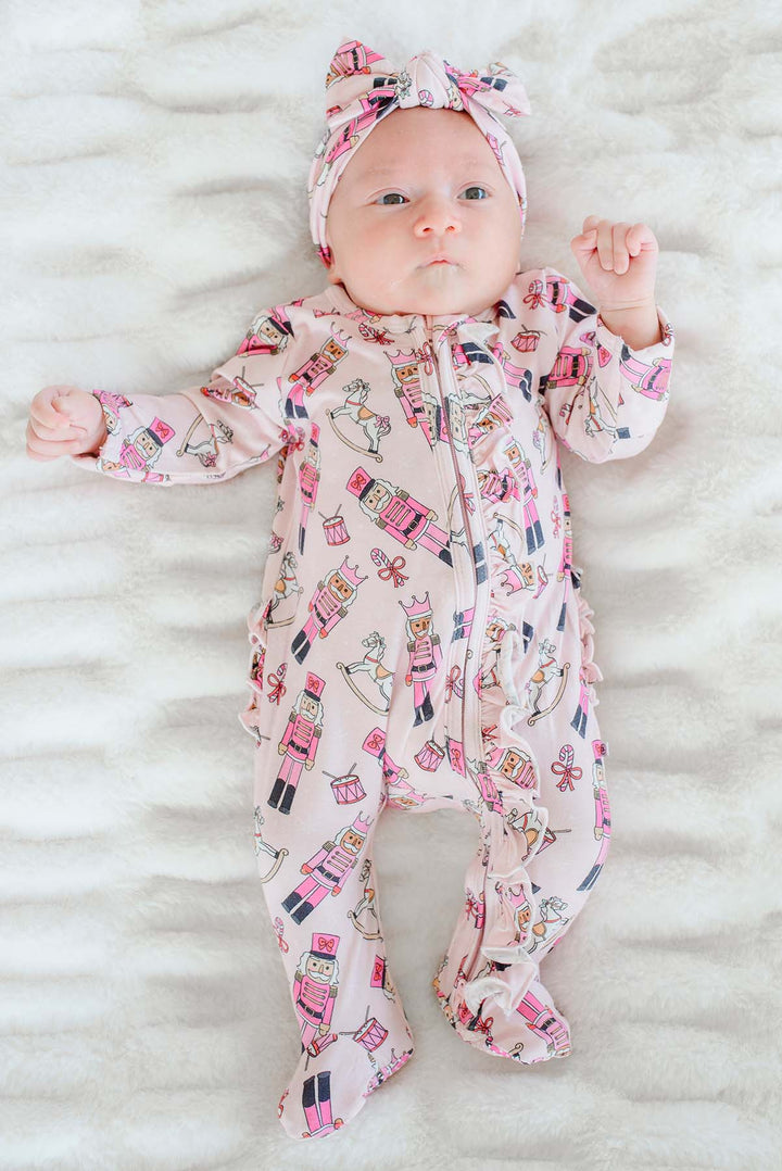 Newborn girl lying down while wearing Caden Lane's pink toy soldier print ruffle zip-up pajamas, featuring nutcrackers and rocking horses, with a matching headband.