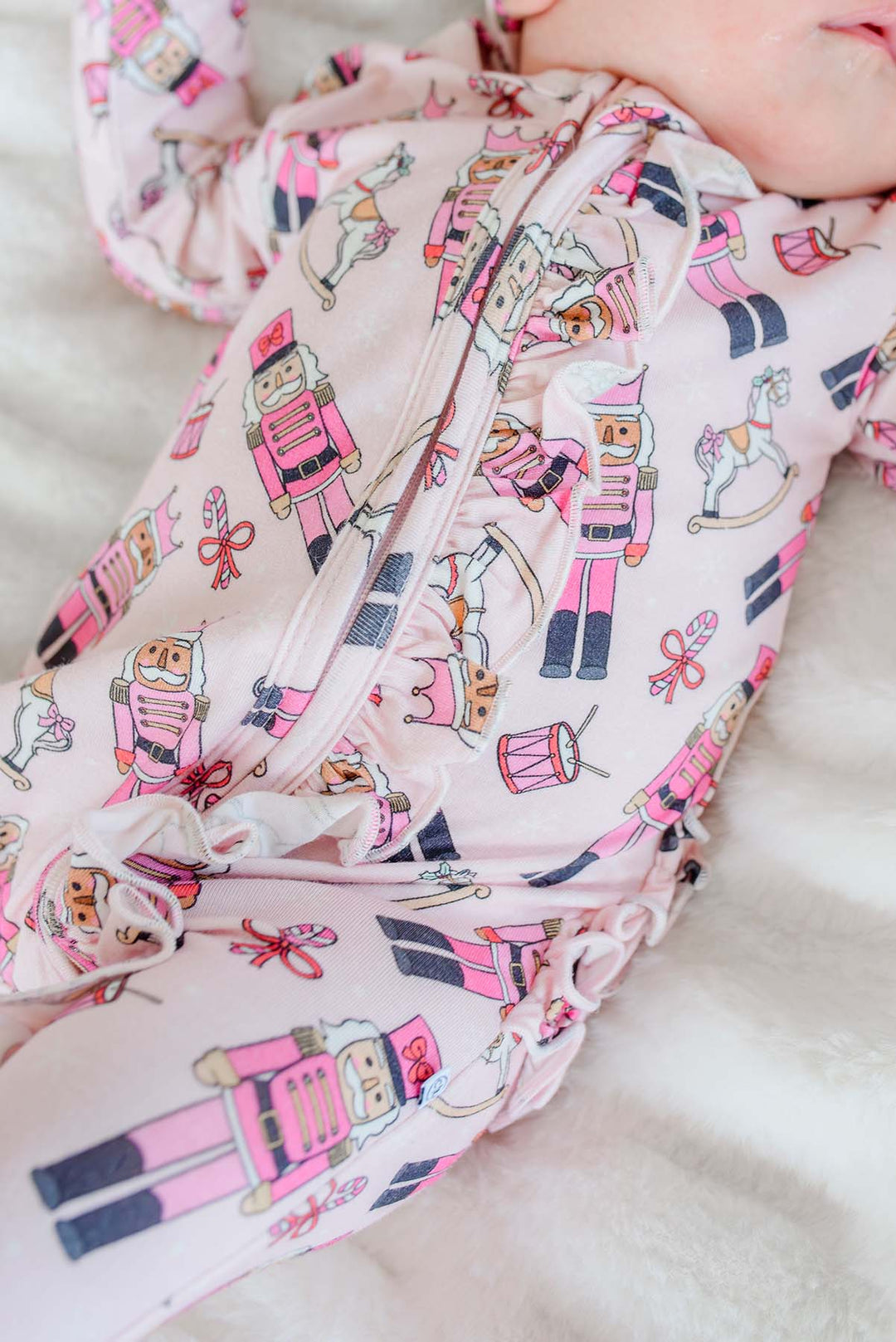 Close-up of Caden Lane's pink toy soldier print ruffle zip-up pajamas, featuring nutcrackers and rocking horses, on a sleeping newborn.