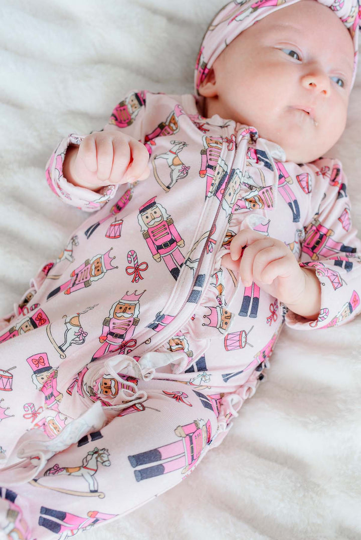 Newborn girl wearing Caden Lane's pink toy soldier print ruffle zip-up pajamas, featuring nutcrackers and rocking horses, with a matching headband.