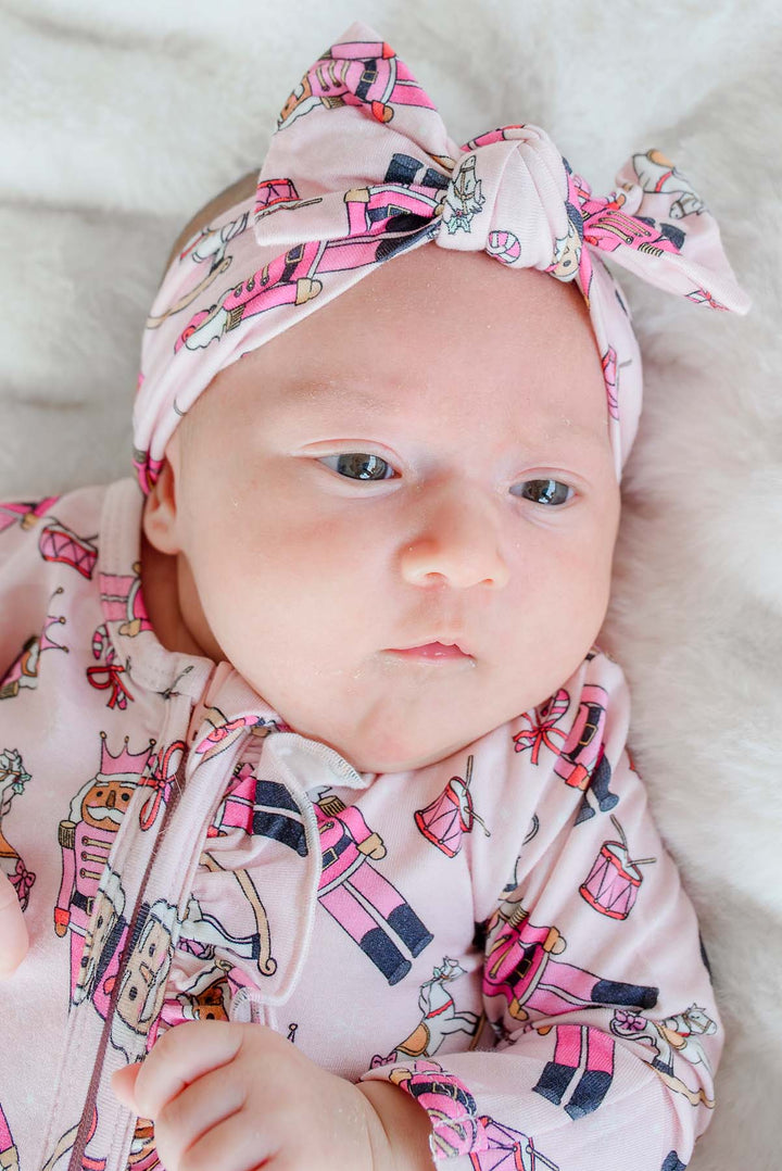 Close-up of newborn girl wearing Caden Lane's pink toy soldier print knot headband and matching zip-up pajamas, featuring nutcrackers and drums.