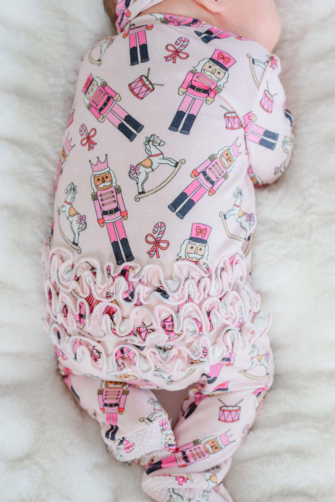 Close-up of newborn girl wearing Caden Lane's pink toy soldier print ruffle zip-up pajamas, featuring nutcrackers and rocking horses.