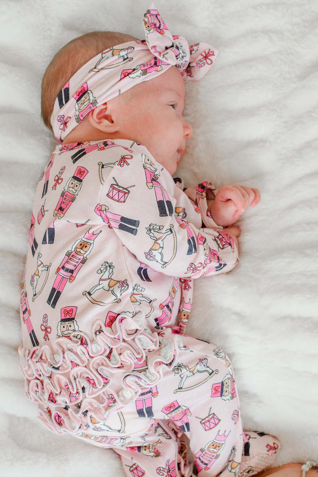 Newborn girl sleeping while wearing Caden Lane's pink toy soldier print ruffle zip-up pajamas, featuring nutcrackers and rocking horses, with a matching headband.