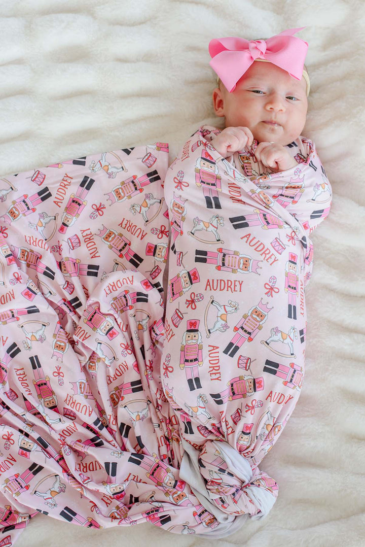 Newborn girl swaddled in Caden Lane's pink toy soldier print blanket personalized with the name "Audrey," featuring nutcrackers, rocking horses, and candy canes, with a pink bow headband.