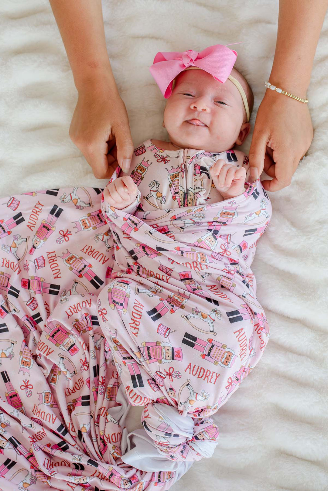 Newborn girl swaddled in Caden Lane's pink toy soldier print blanket personalized with the name "Audrey," featuring nutcrackers, rocking horses, and candy canes, wearing a pink bow headband.