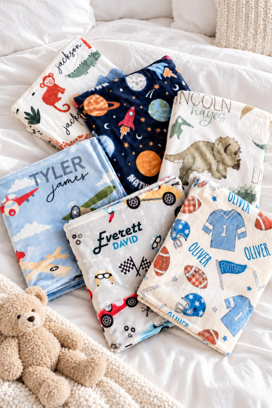Six Caden Lane personalized themed blankets with various colorful cartoon and sports-themed prints are arranged on a white bed alongside a plush teddy bear.
