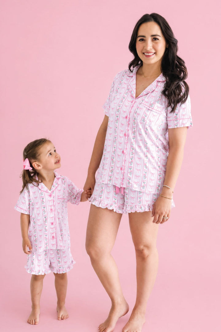 A woman and a young girl wearing matching pink and white patterned pajamas, standing barefoot against a solid pink studio background.