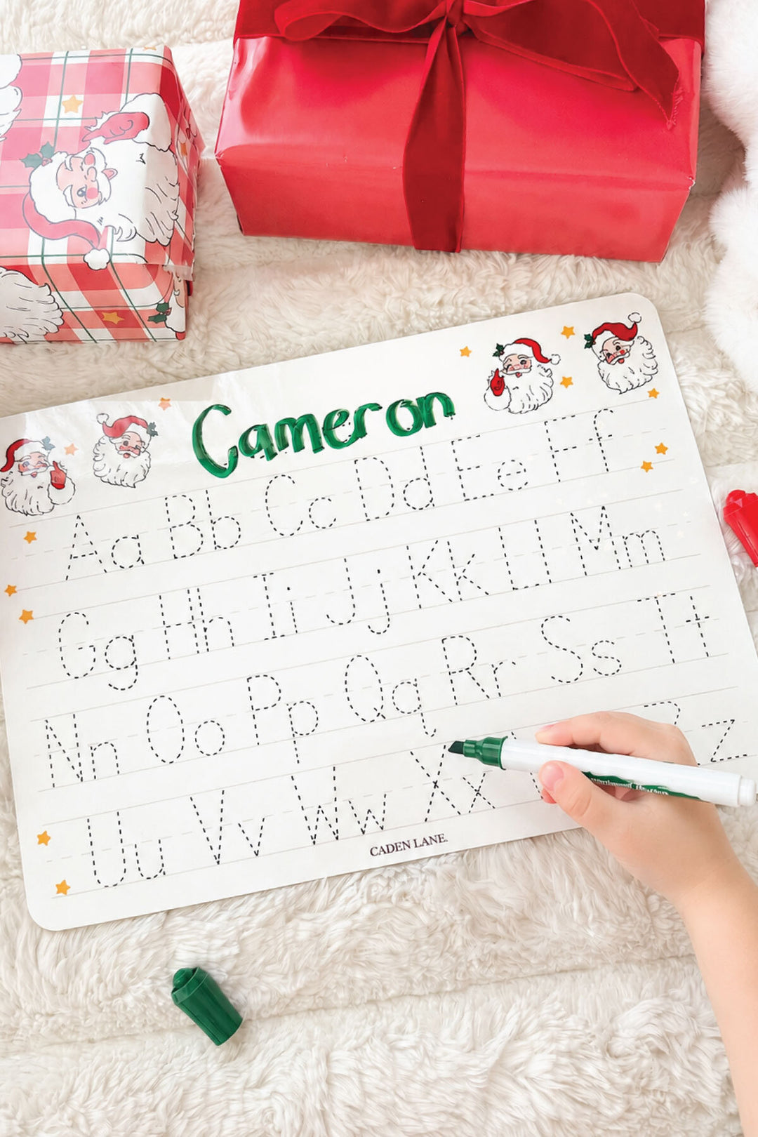 Caden Lane personalized Christmas handwriting practice board featuring the name "Cameron" in festive red and green plaid lettering with traceable uppercase and lowercase alphabet, creating a fun personalized holiday learning gift for kids.