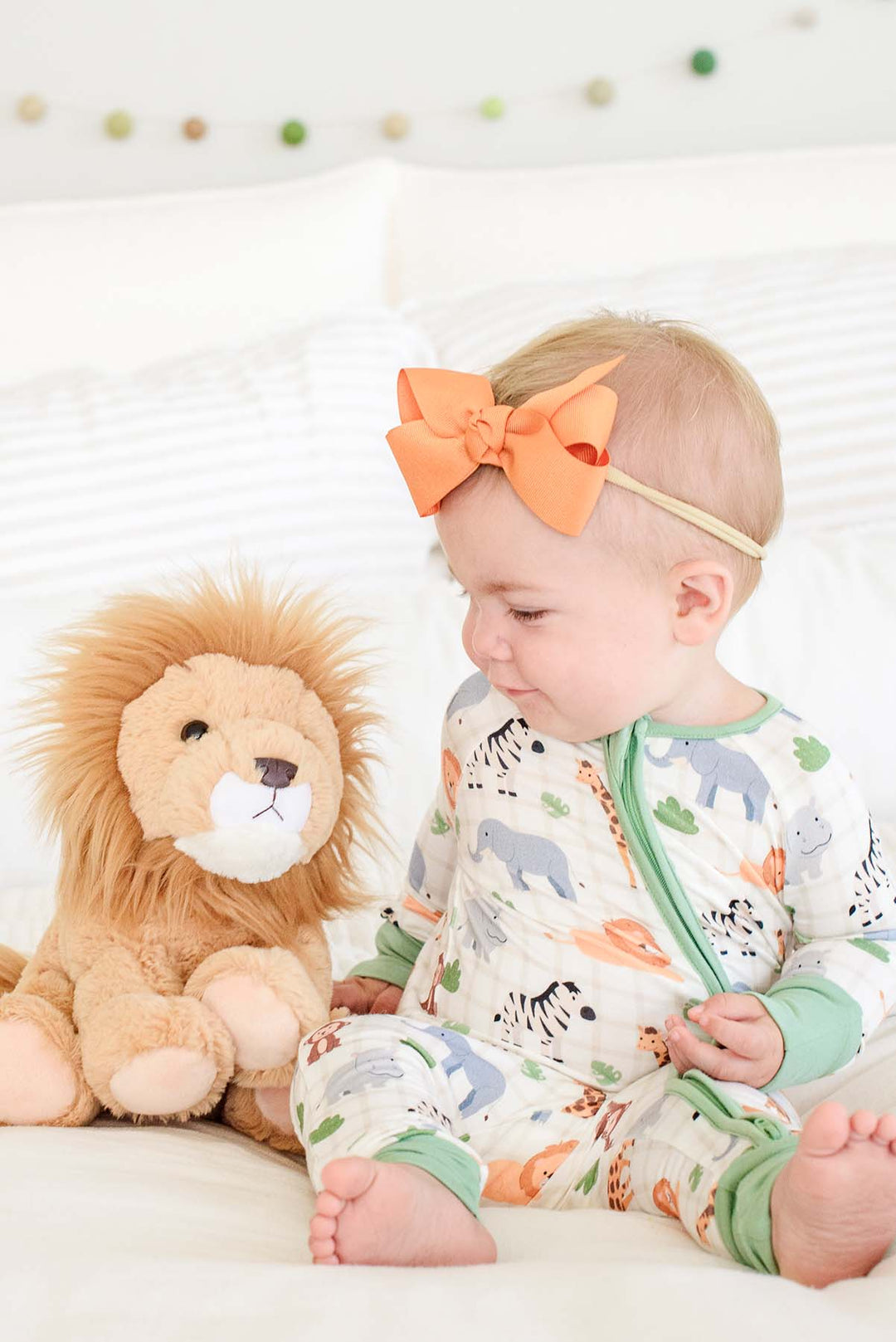 Baby girl sitting on a bed and smiling at a lion plush, wearing a Caden Lane Wild Child convertible zip romper with green trim, paired with an orange bow headband.