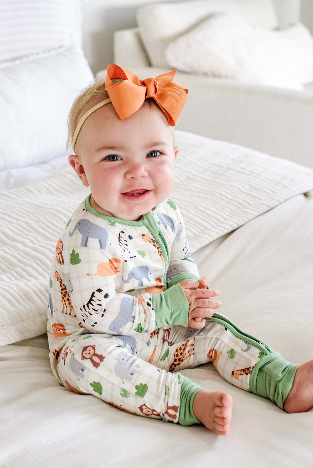 Smiling baby girl sitting on a white bed in a Caden Lane convertible zip romper in a Wild Child print featuring elephants, zebras and giraffes, paired with an orange bow headband.