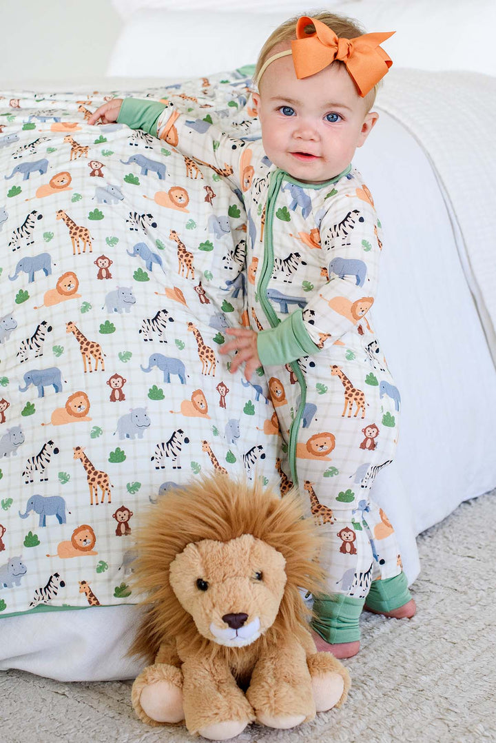 Baby girl standing beside a bed draped in a Caden Lane Wild Child bamboo blanket, paired with a matching zip romper with green trim, an orange bow headband and a lion plush.

