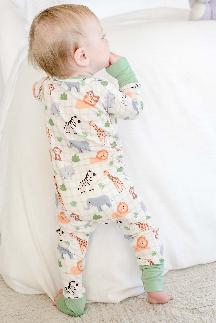 Back view of a baby boy standing against a bed with his hand to his mouth, wearing a Caden Lane Wild Child convertible zip romper with green cuffs.