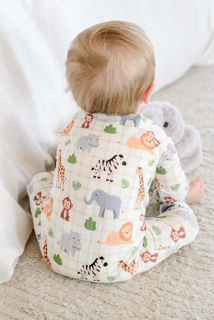 Back view of a baby boy sitting on a rug beside an elephant plush, wearing a Caden Lane convertible zip romper in a Wild Child print.