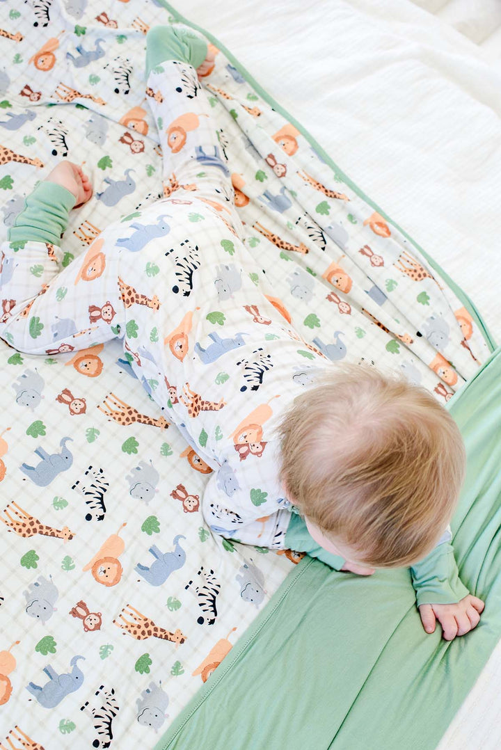 Overhead view of a baby boy crawling across a Caden Lane bamboo blanket in a Wild Child print with sage green trim, paired with a matching zip romper.