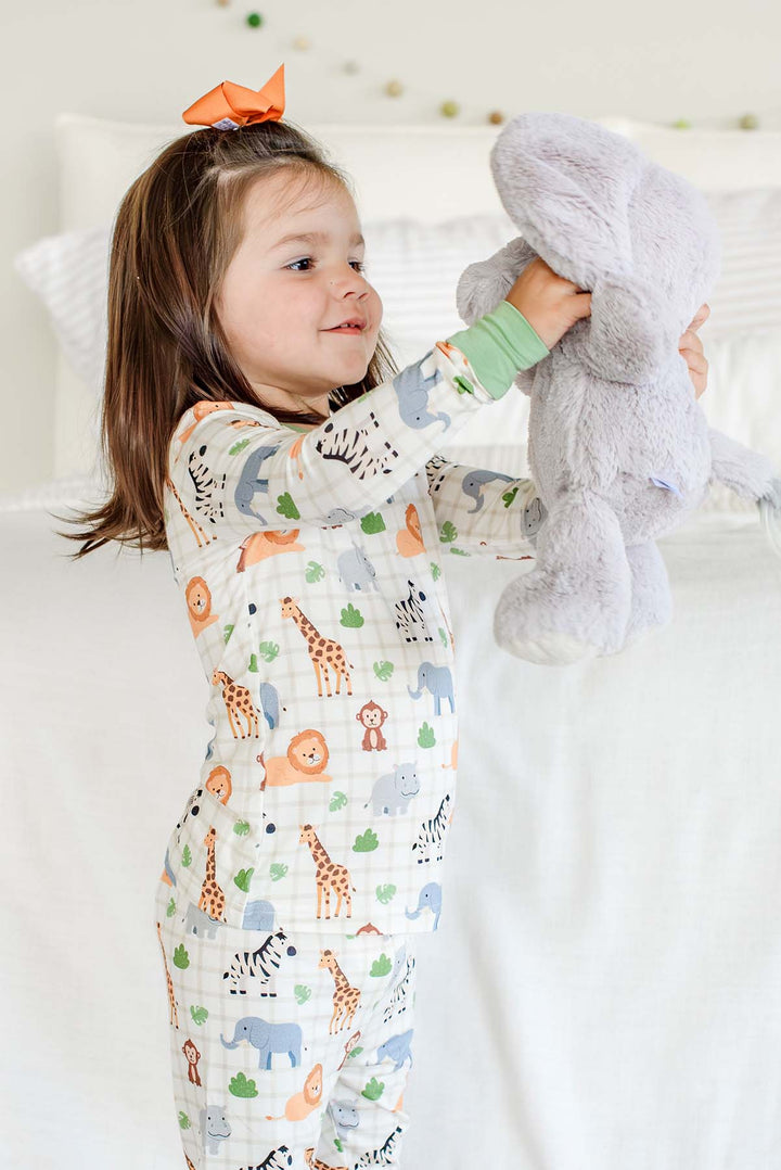 Girl holding up a gray elephant plush while wearing a Caden Lane two-piece pajama set in a Wild Child print with green cuffs, paired with an orange hair bow.