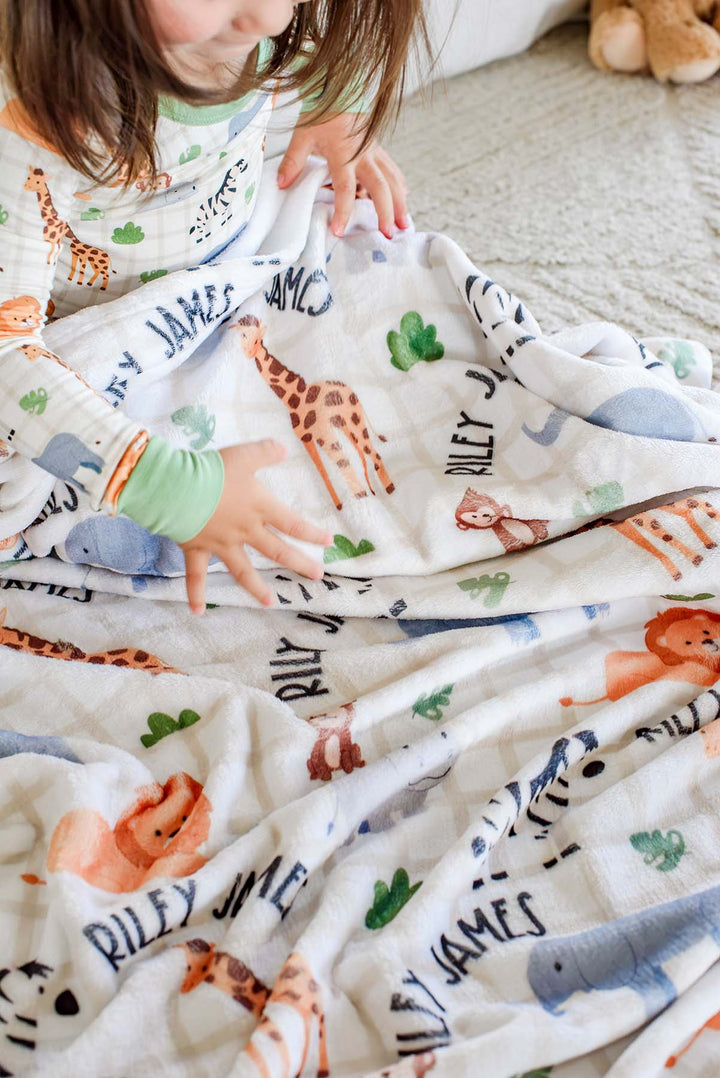 Toddler girl in matching Wild Child pajamas spreading out a Caden Lane personalized plush blanket reading "Riley James" across a cream rug.