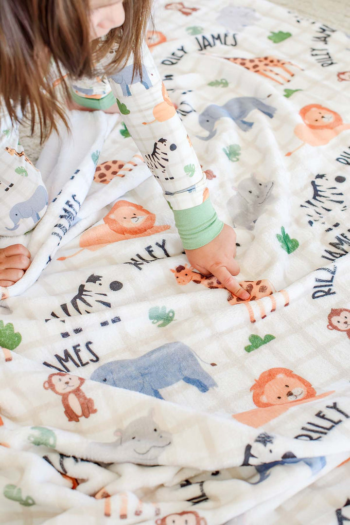 Close-up of a toddler's finger pointing to a giraffe on a Caden Lane personalized Wild Child plush blanket reading "Riley James," paired with matching pajamas with green cuffs.