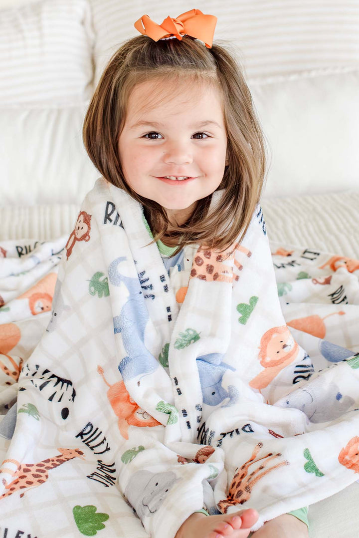 Smiling toddler girl wrapped in a Caden Lane personalized plush blanket in a Wild Child print reading "Riley James," paired with an orange hair bow.
