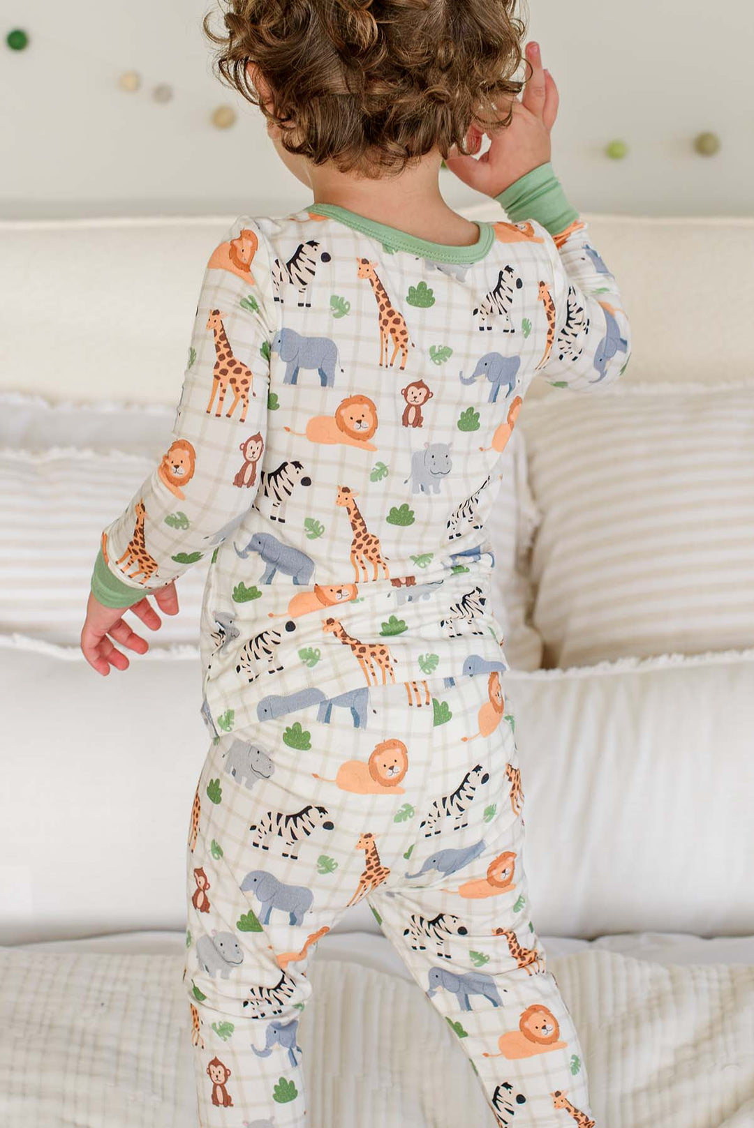 Back view of a curly-haired boy standing on a bed in a Caden Lane two-piece pajama set in a Wild Child print featuring giraffes, lions, zebras and elephants.