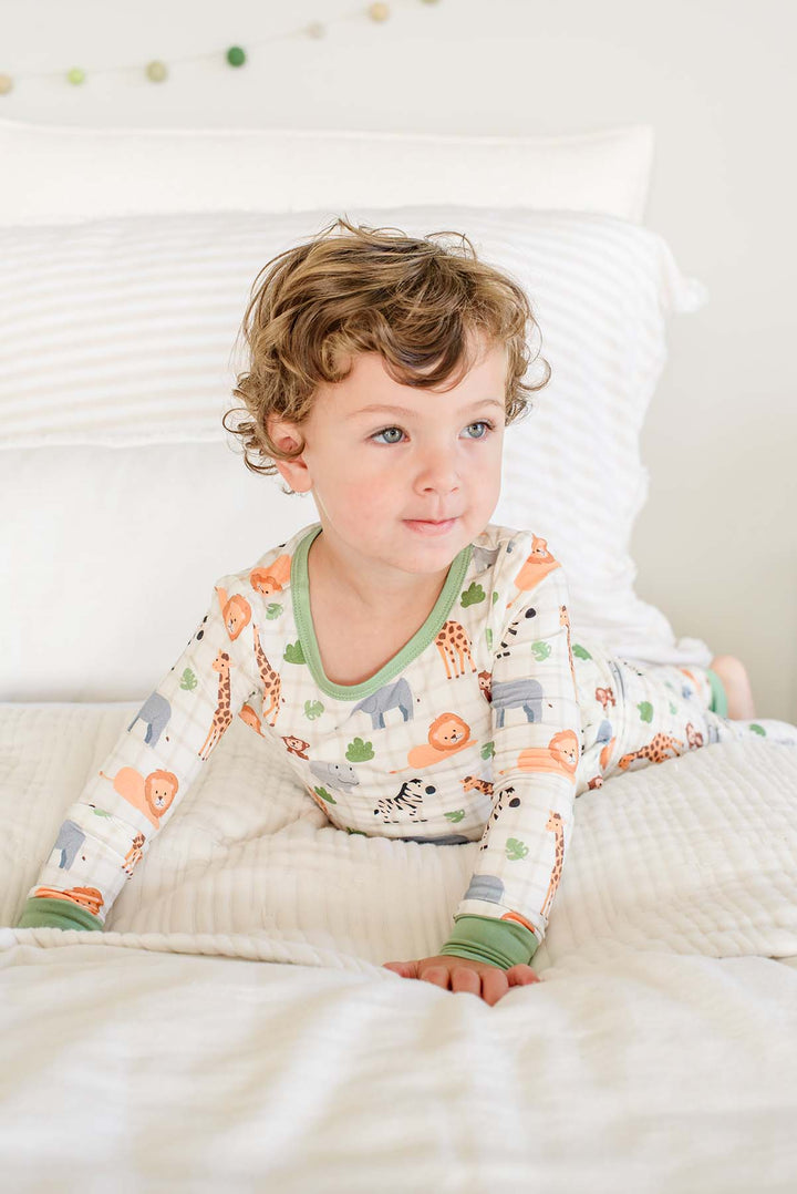 Curly-haired boy lying on his stomach on a white bed, wearing a Caden Lane two-piece pajama set in a Wild Child print with green neckline and cuffs.