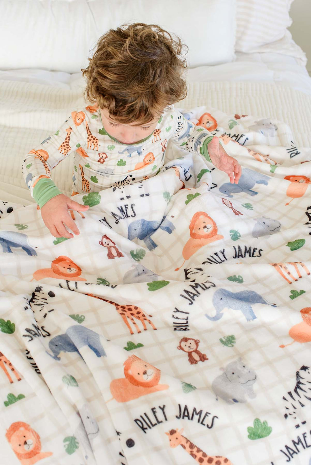 Overhead view of a toddler boy in matching Wild Child pajamas reaching across a Caden Lane personalized plush blanket reading "Riley James" spread over a bed.