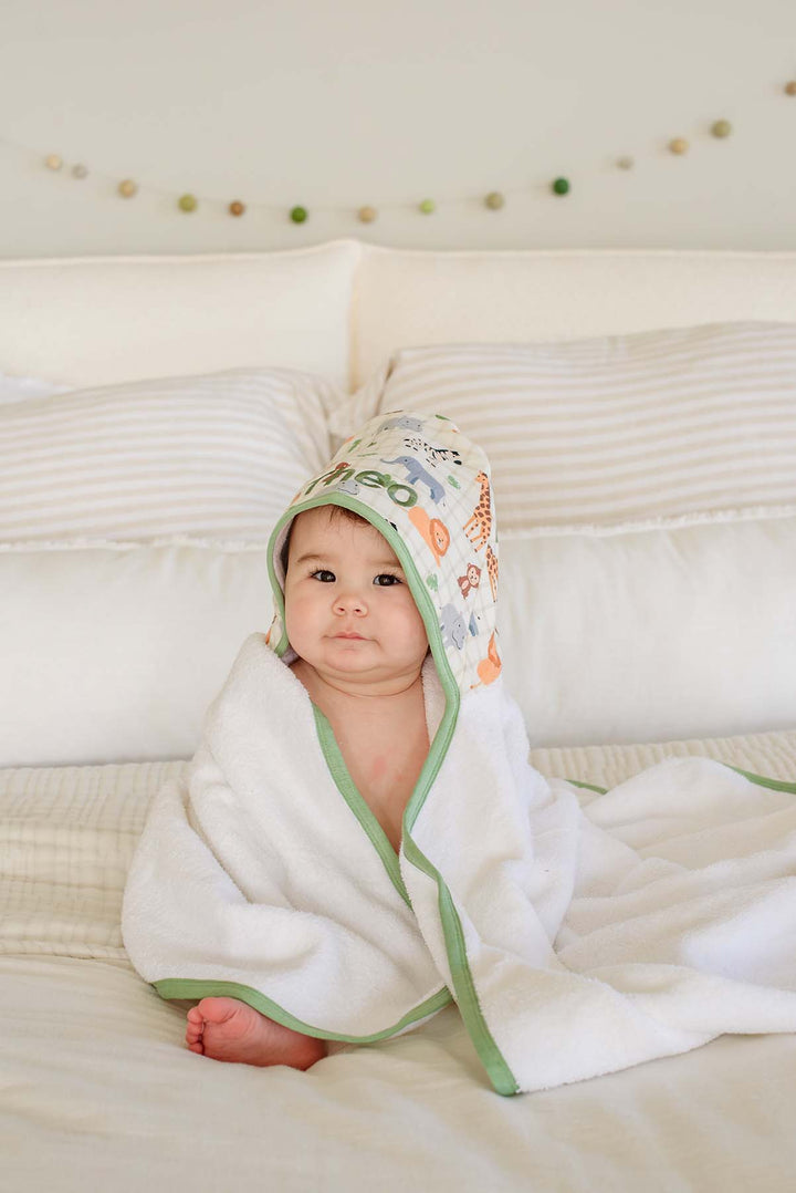 Baby sitting on a bed against striped pillows, wrapped in a Caden Lane hooded towel with a Wild Child print hood reading "Theo," with a felt ball garland above.