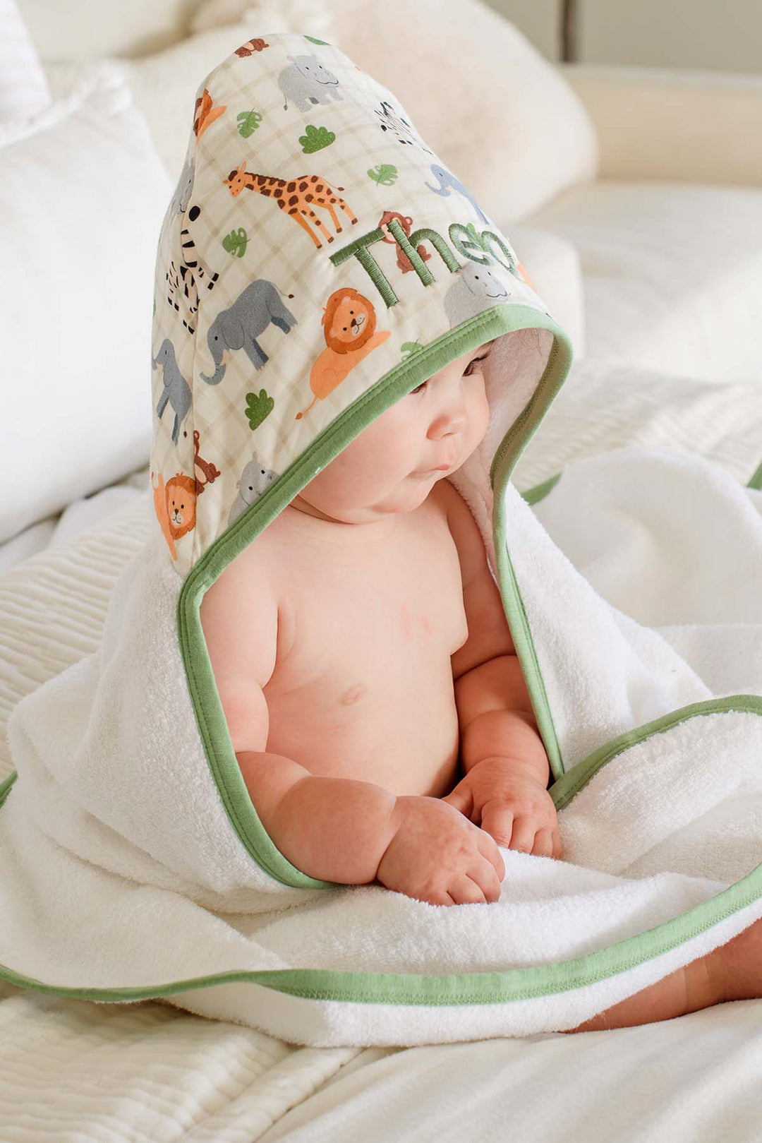 Side view of a baby looking away while wrapped in a Caden Lane hooded towel, showing the Wild Child print hood with giraffes, lions and elephants embroidered with "Theo."