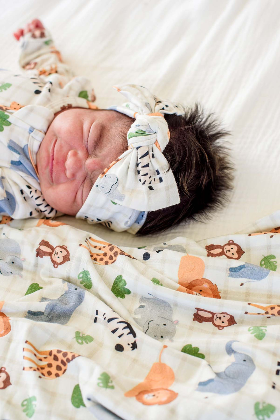 Close-up of a sleeping newborn wearing a Caden Lane knotted bow headwrap in a Wild Child print, lying on a matching blanket with lions, giraffes and monkeys.