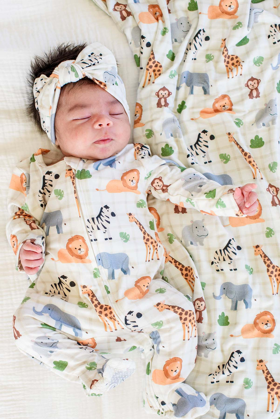 Sleeping newborn wearing a Caden Lane zipper footie in a Wild Child print featuring lions, zebras, giraffes and elephants, paired with a matching knotted bow headwrap and blanket.