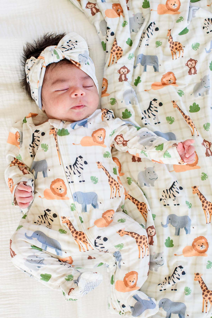 Sleeping newborn wearing a Caden Lane zipper footie in a Wild Child print featuring lions, zebras, giraffes and elephants, paired with a matching knotted bow headwrap and blanket.