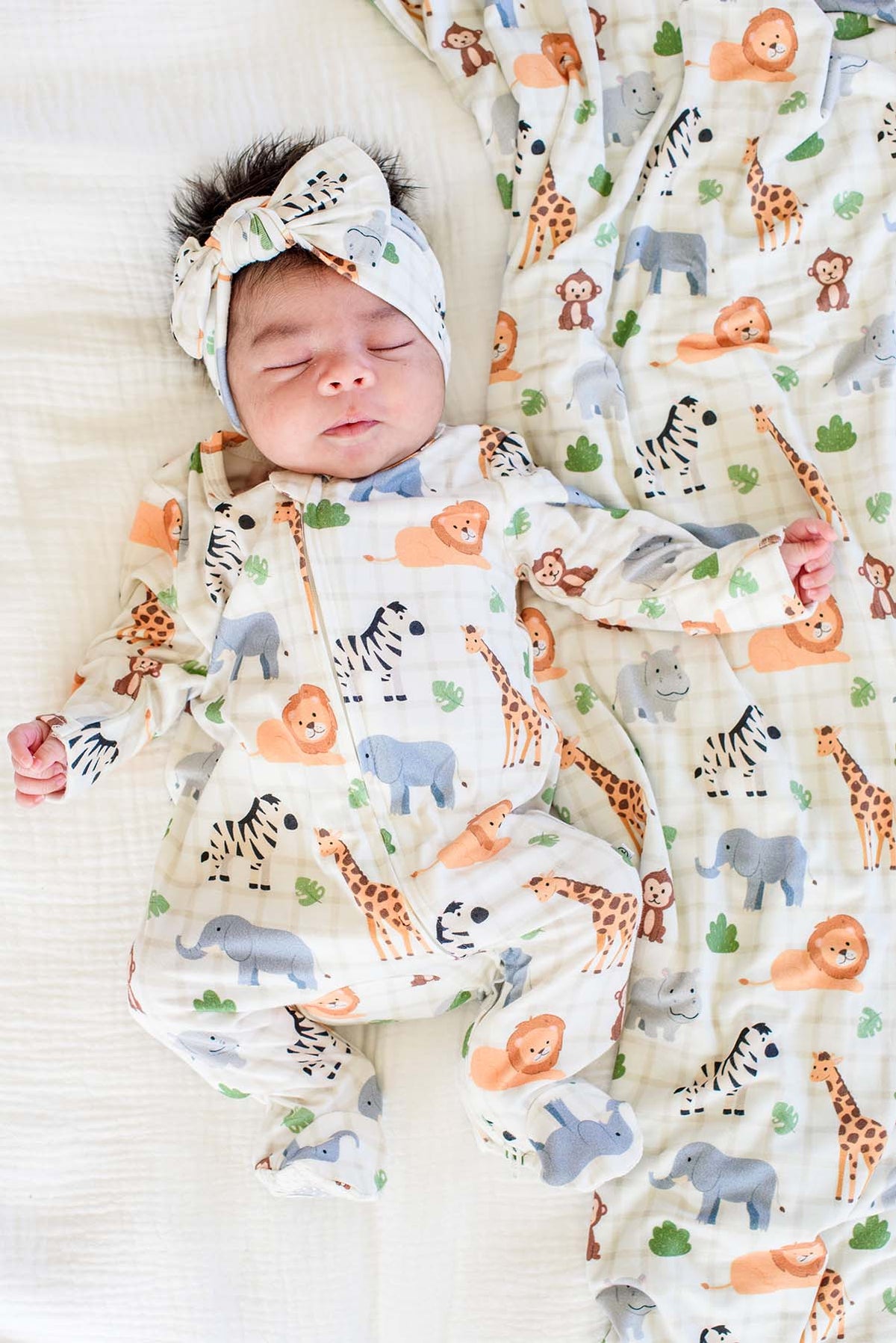 Overhead view of a sleeping newborn in a Caden Lane zipper footie in a Wild Child print, paired with a matching knotted bow headwrap and blanket.