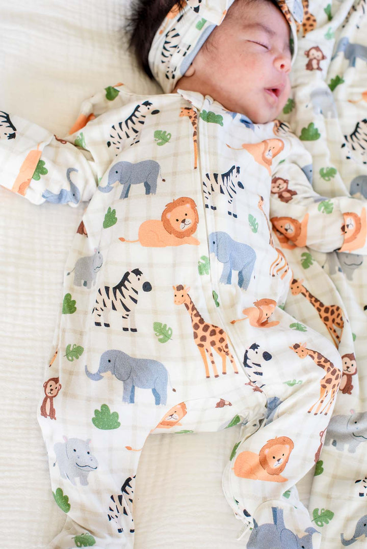 Close-up of a sleeping newborn in a Caden Lane zipper footie in a Wild Child print featuring zebras, elephants, lions and giraffes, paired with a matching headwrap.