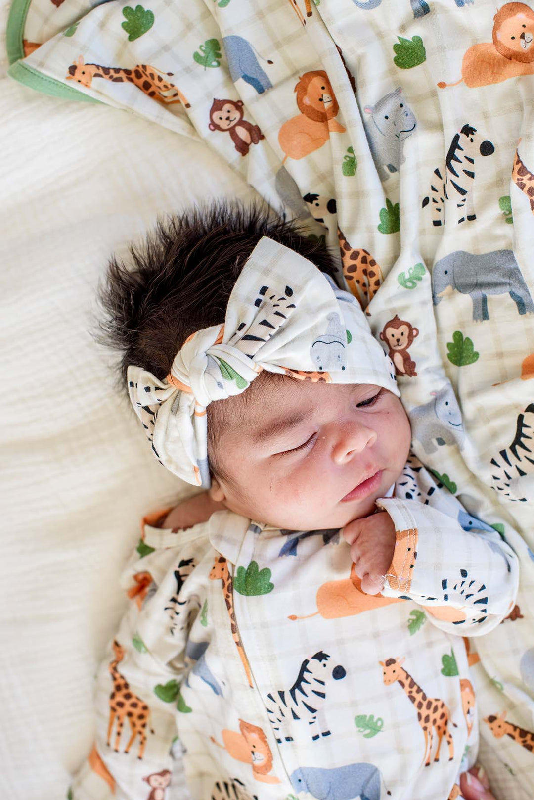 Overhead close-up of a sleeping newborn wearing a Caden Lane Wild Child knotted bow headwrap and matching footie, resting on a Wild Child blanket with green trim.