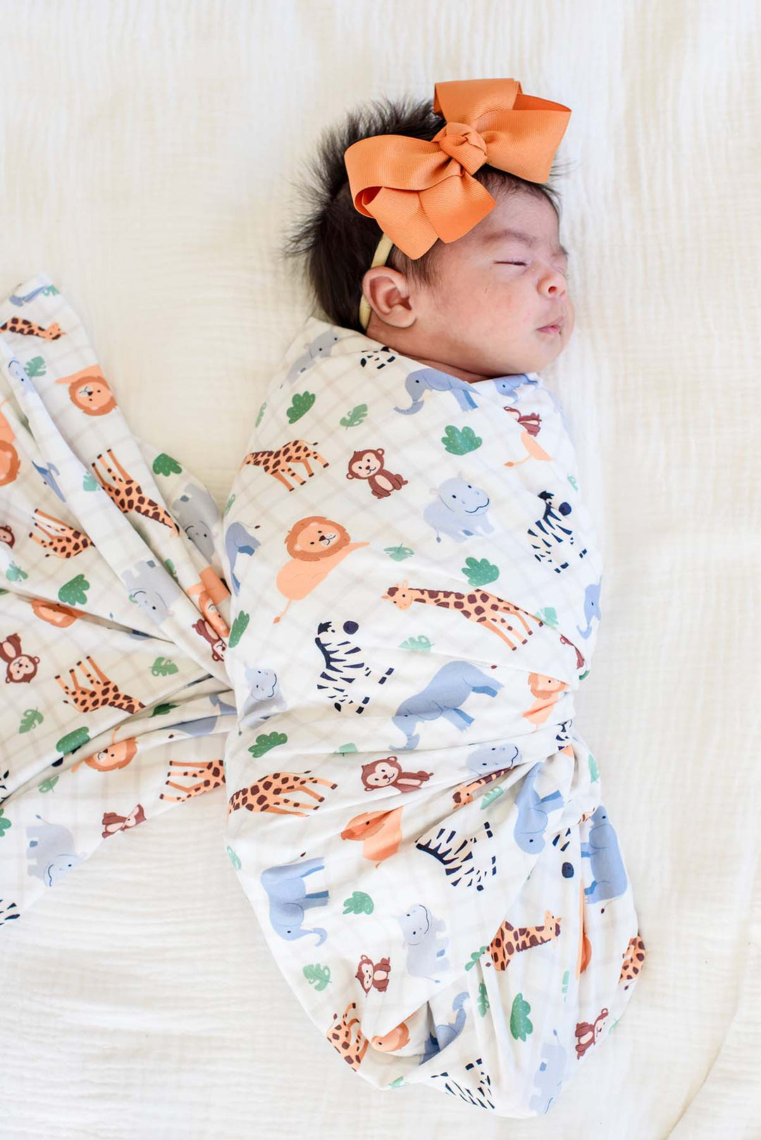 Sleeping newborn swaddled in a Caden Lane swaddle blanket in a Wild Child print featuring safari animals and green leaves on cream plaid, paired with an orange bow headband.