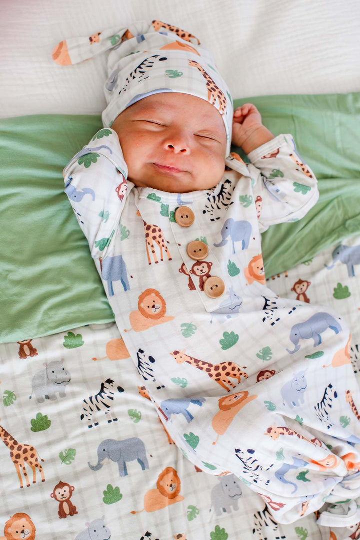 Close-up of a sleeping newborn stretching in a Caden Lane knotted gown with wooden buttons and a matching knot hat in a Wild Child print featuring giraffes, monkeys and lions.