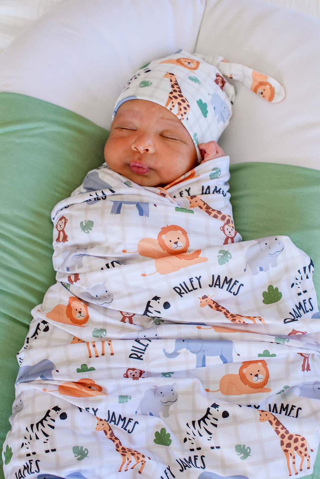 Sleeping newborn swaddled in a Caden Lane personalized swaddle blanket in a Wild Child print reading "Riley James," paired with a matching knot hat and a sage green blanket underneath.