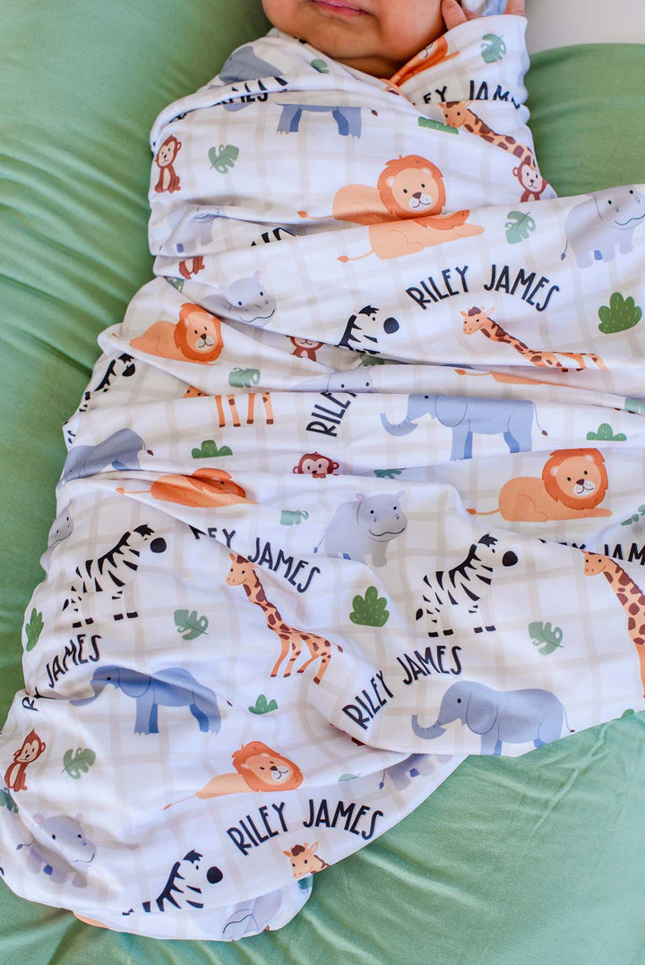 Close-up of a newborn snugly wrapped in a Caden Lane personalized Wild Child swaddle blanket featuring lions, giraffes, zebras and elephants with arched name text reading "Riley James."