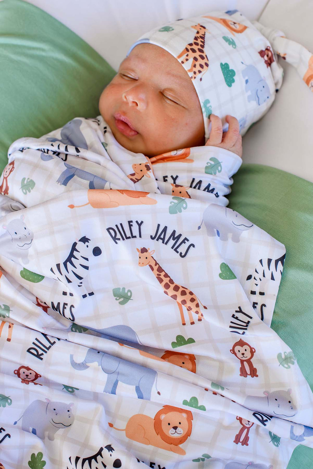 Close-up of a sleeping newborn with a hand by his face, swaddled in a Caden Lane personalized Wild Child swaddle reading "Riley James," paired with a matching knot hat.
